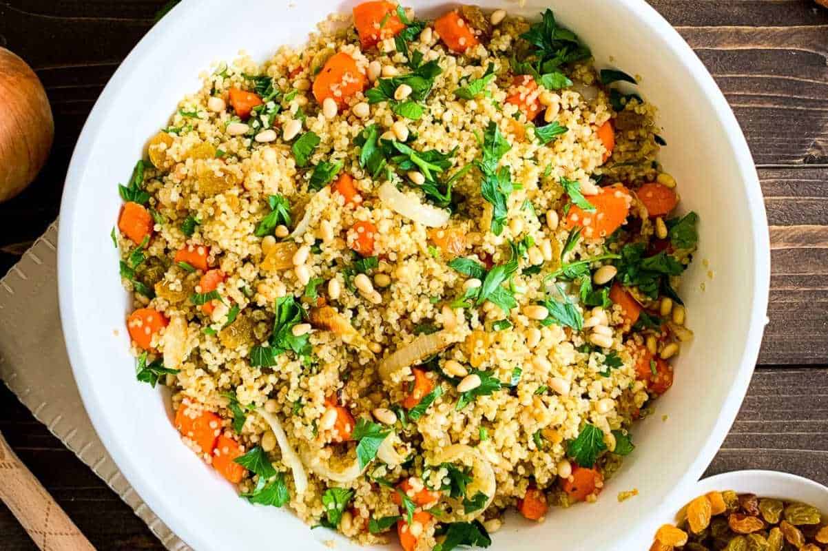 A bowl of quinoa salad with roasted vegetables, pine nuts, and fresh basil, served in a white dish on a wooden table.