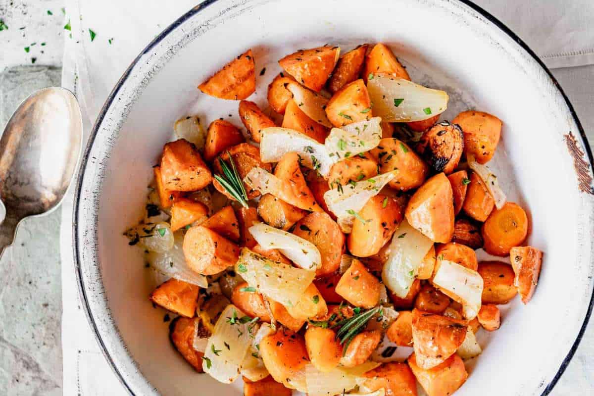 A bowl of roasted sweet potatoes and onions garnished with herbs, accompanied by a spoon on a light surface.