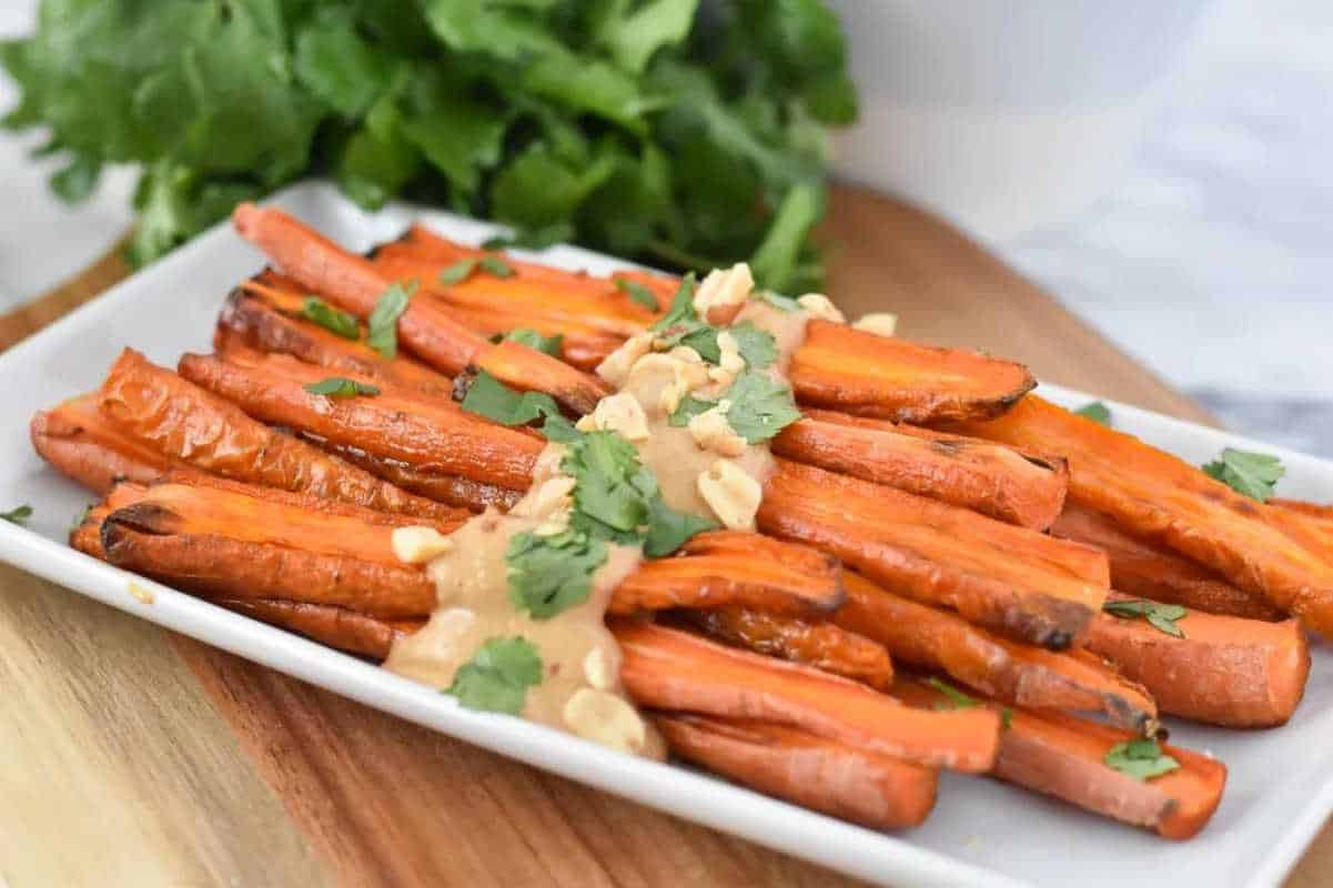 Roasted carrots topped with peanut sauce and garnished with cilantro on a white plate, fresh greens in the background.