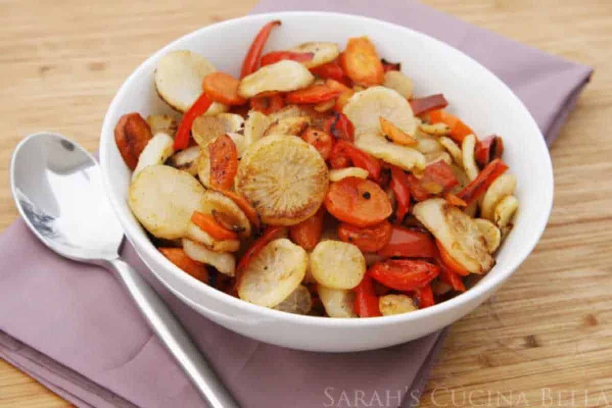 A bowl of roasted vegetables including sliced carrots, red peppers, and parsnips on a wooden table, with a spoon on the side.