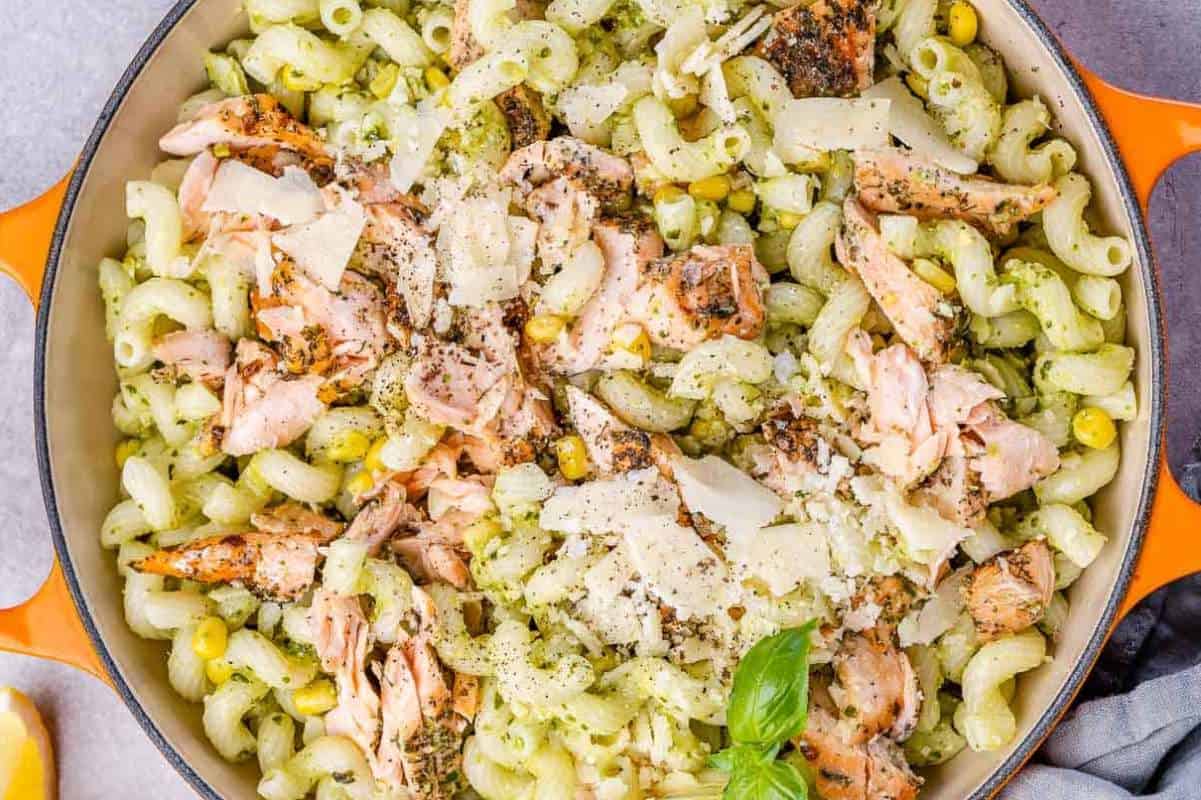 A skillet with pesto pasta, flaked salmon, corn, and grated cheese, garnished with basil leaves.
