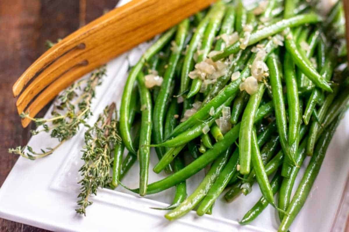 A plate of cooked green beans garnished with herbs and minced garlic, served with a wooden serving spoon.