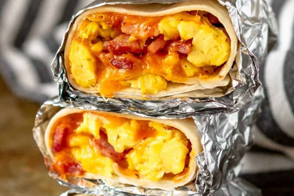 Two halves of a breakfast burrito wrapped in foil, filled with scrambled eggs, bacon, and cheese.