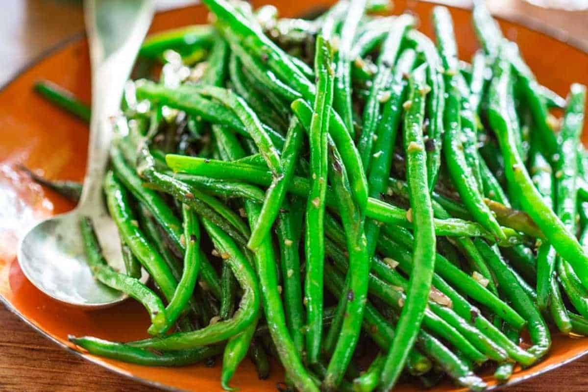 A plate of sautéed green beans garnished with spices, served with a spoon on a wooden table.