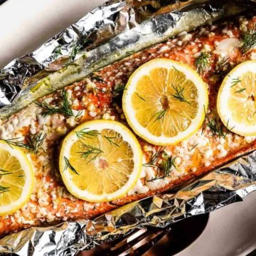 Baked trout topped with lemon slices and dill, wrapped in foil on a white surface.