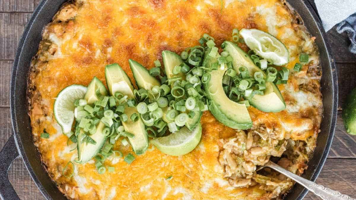 A skillet with enchilada casserole topped with melted cheese, avocado slices, lime, and chopped green onions.