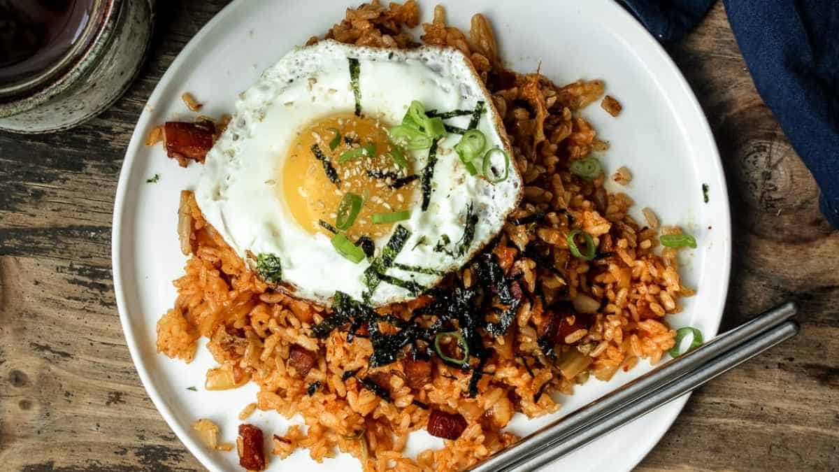 A plate of kimchi fried rice topped with a fried egg and garnished with green onions and seaweed strips, accompanied by a pair of chopsticks.