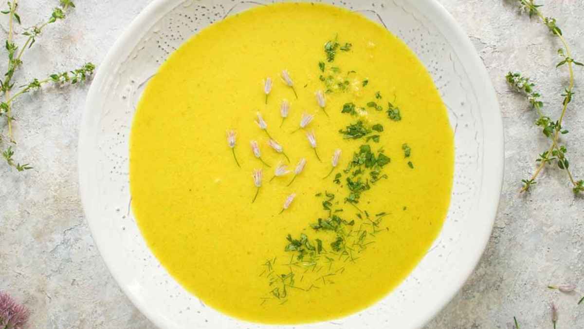 A bowl of creamy yellow soup garnished with herbs and small purple flowers, placed on a textured grey surface.