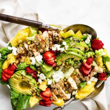 A fresh salad with grilled chicken, avocado, strawberries, and crumbled cheese served in a metallic bowl.