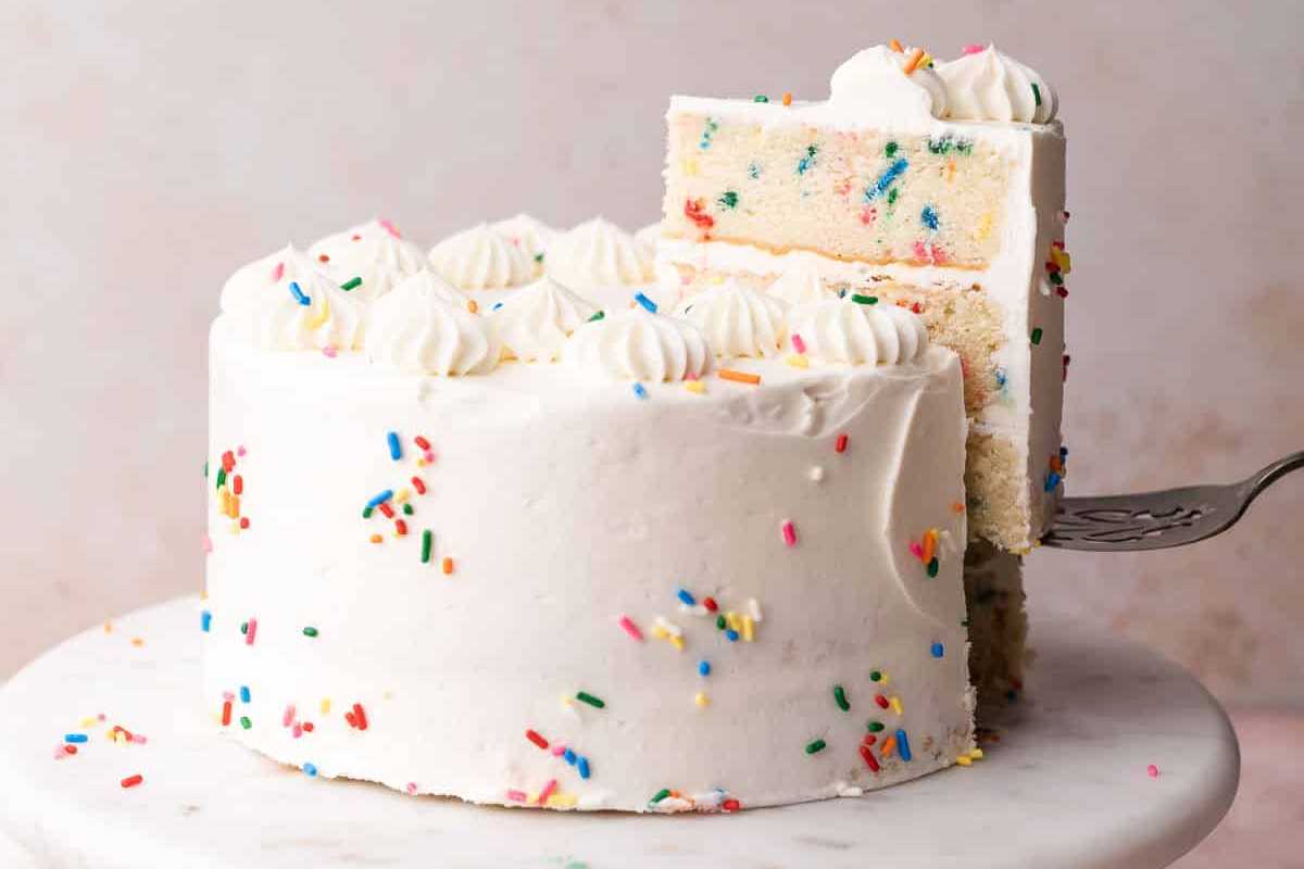 A vanilla cake with white frosting and colorful sprinkles, with one slice being lifted by a cake server.