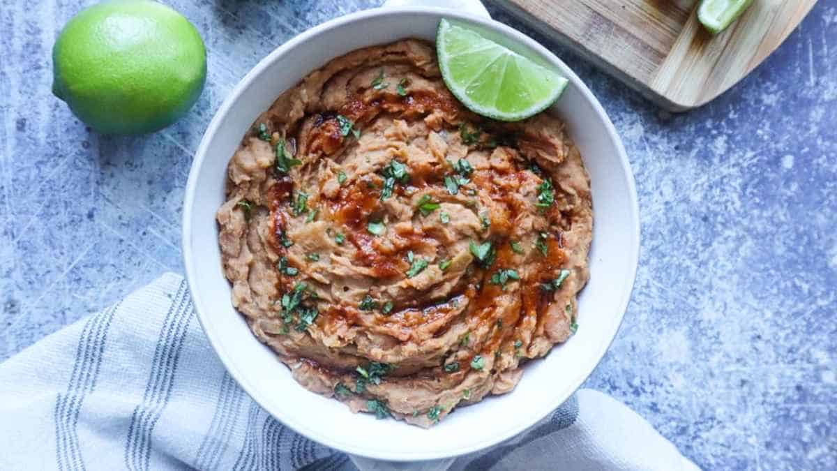 A bowl of creamy refried beans garnished with chopped cilantro and a drizzle of sauce, accompanied by a cut lime on a blue textured surface.