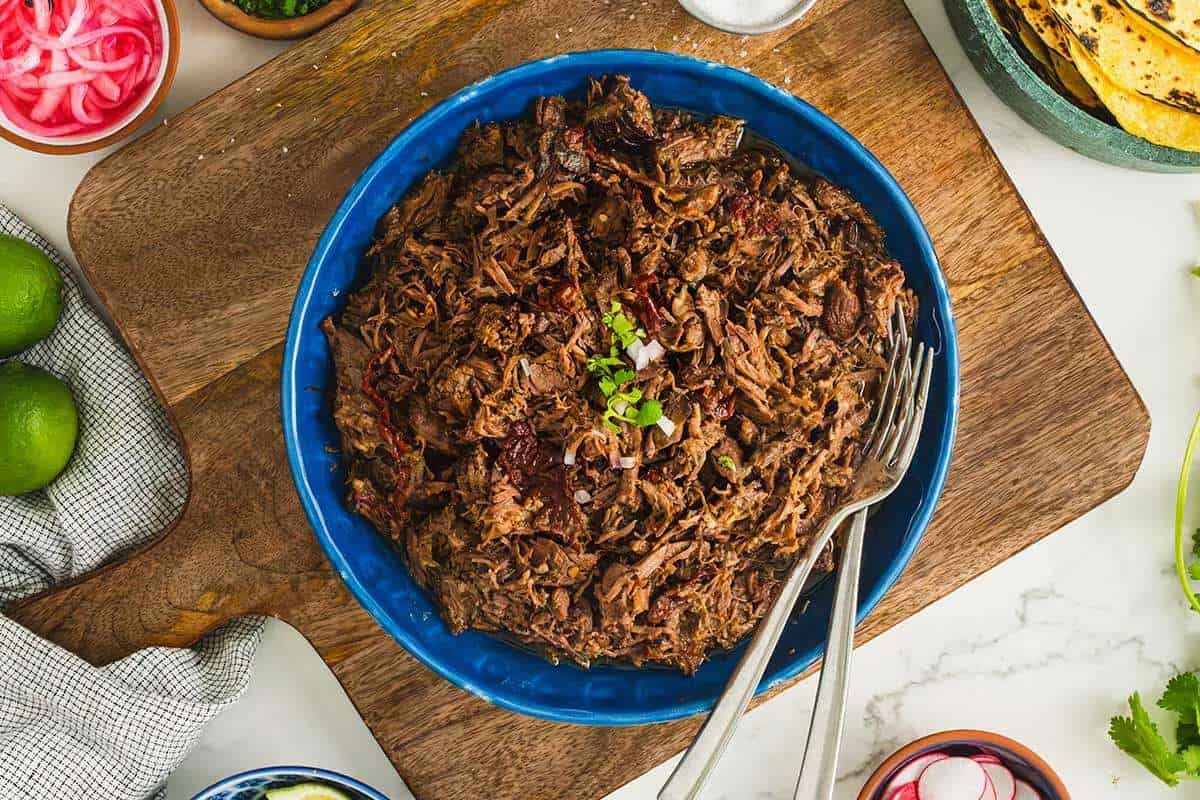 A blue bowl filled with shredded beef on a wooden board, accompanied by lime wedges and tortillas, with pickled onions on the side.