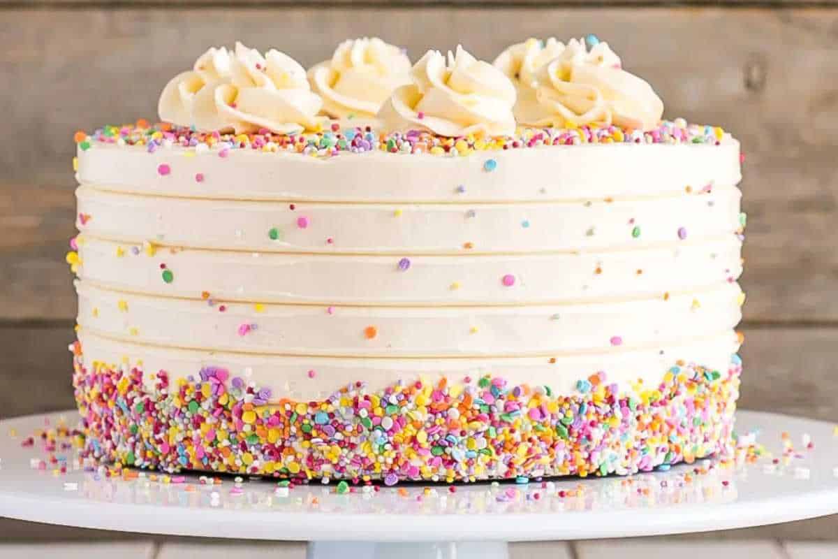 A layered vanilla cake with white frosting and colorful sprinkles on its sides, presented on a white plate.