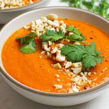 A bowl of creamy carrot soup garnished with chopped nuts and fresh cilantro, served on a light-colored surface.
