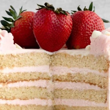 A strawberry cake with pink frosting and fresh strawberries on top, displayed with one slice cut out to reveal layered interior.