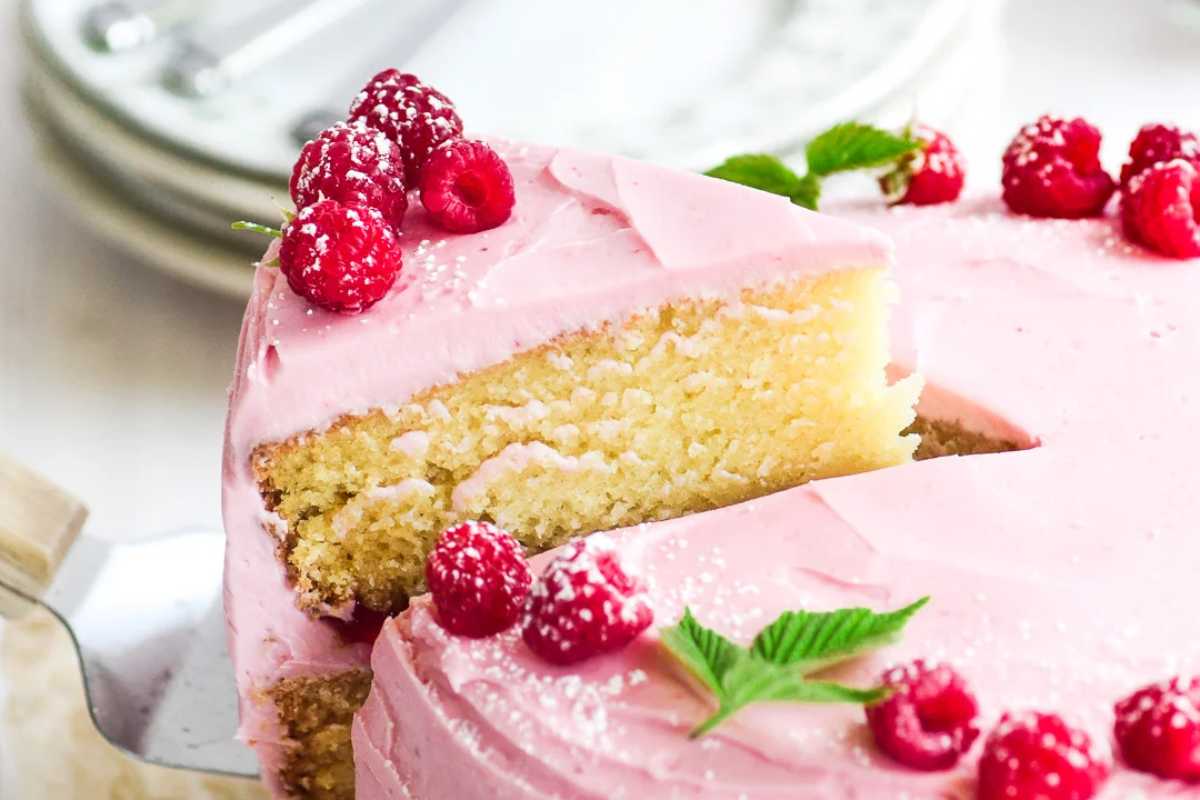 A slice of raspberry cake with pink frosting and fresh raspberries on top, garnished with mint leaves, on a white plate.