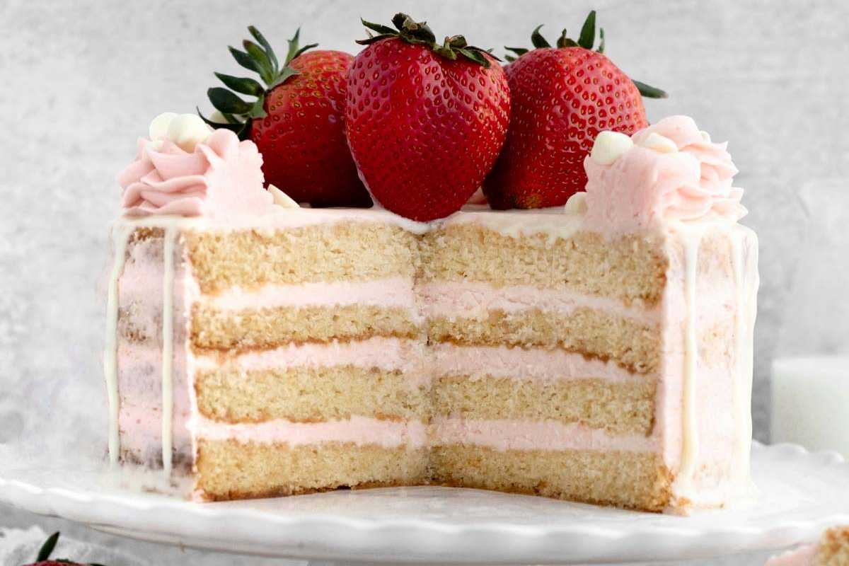 A sliced strawberry layer cake with pink frosting and whole strawberries on top, displayed on a white plate.