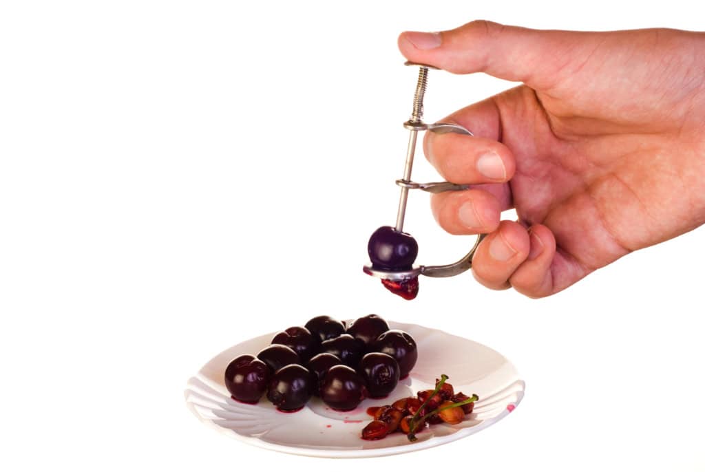 A hand using a cherry pitter to remove pits from cherries over a plate with pitted cherries and pits.
