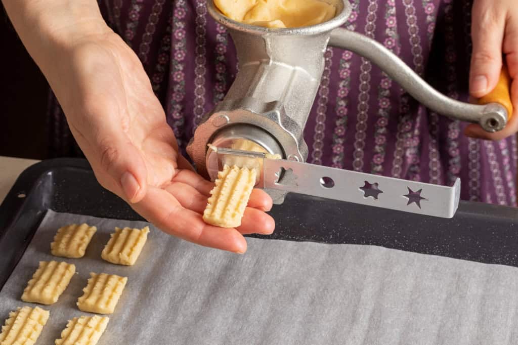 A person using a cookie press to extrude dough onto a baking sheet, forming small patterned cookies.