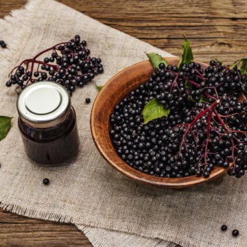A wooden bowl full of elderberries on a rustic table, with a small jar of elderberry jam and scattered berries around.