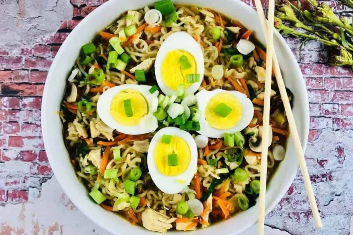A bowl of ramen noodles topped with sliced boiled eggs, green onions, and mixed vegetables, served with chopsticks on the side.