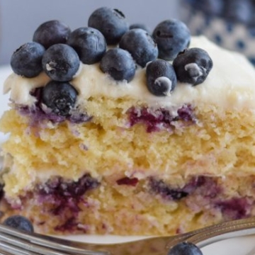 A slice of blueberry cake with fresh blueberries on top, served on a white plate with a fork on the side.