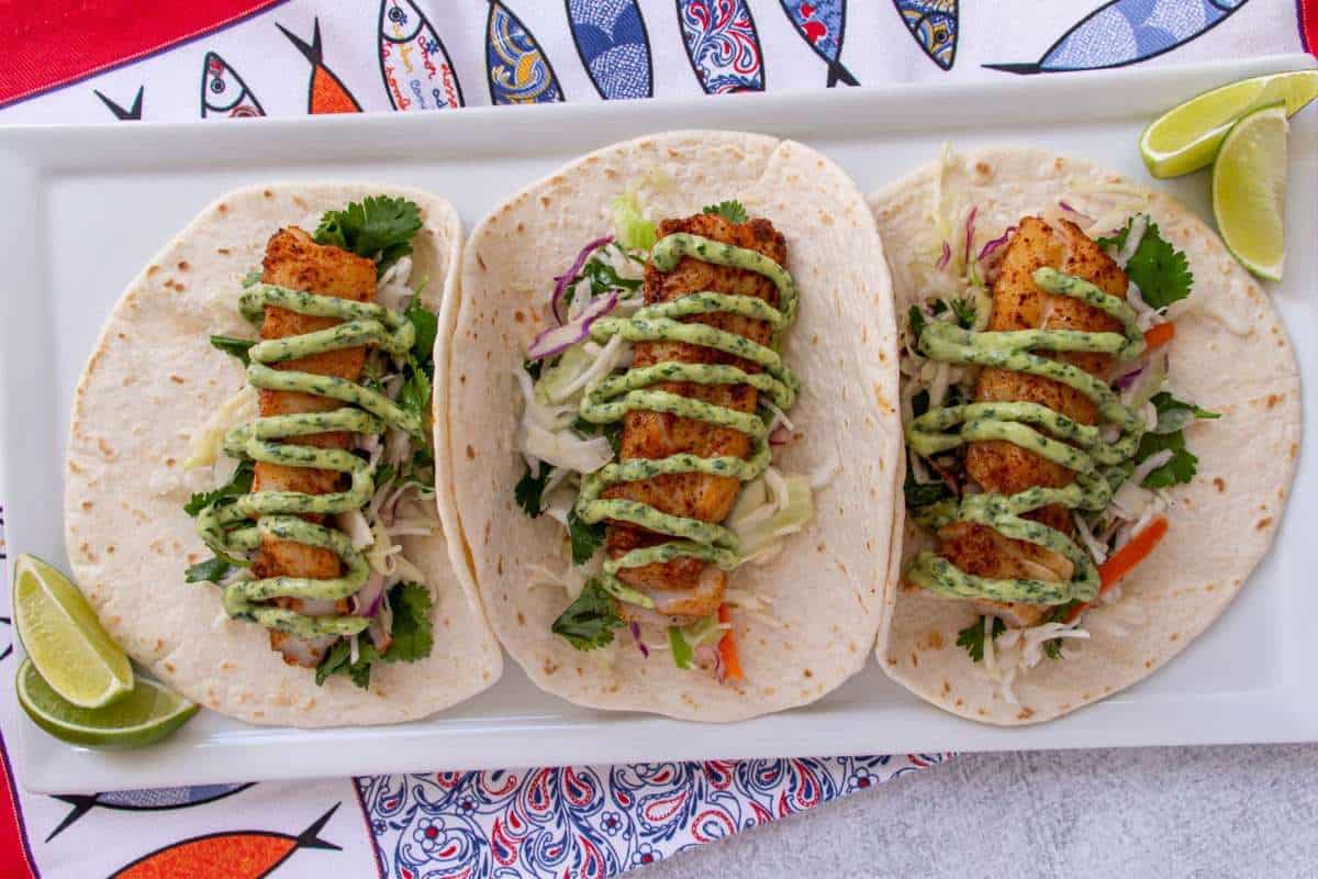 Three fish tacos with cabbage slaw and avocado sauce on tortillas, garnished with lime wedges, served on a colorful fish-themed plate.