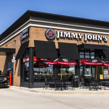 Exterior view of a Jimmy John's gluten-free restaurant with black umbrellas and outdoor seating on a sunny day.