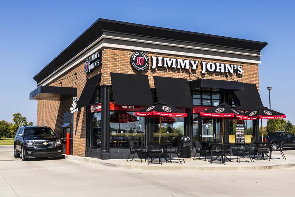 Exterior view of a Jimmy John's gluten-free restaurant with black umbrellas and outdoor seating on a sunny day.