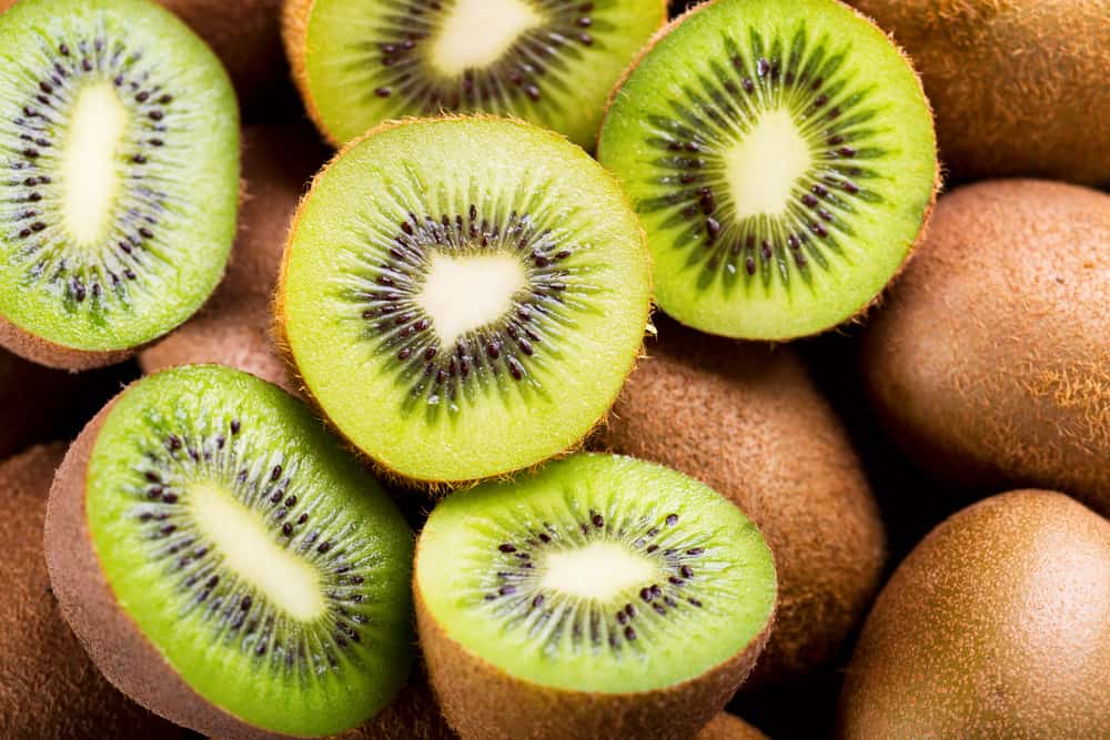 Sliced kiwi fruits displaying vibrant green flesh with black seeds, arranged among whole, brown, fuzzy kiwi skins.