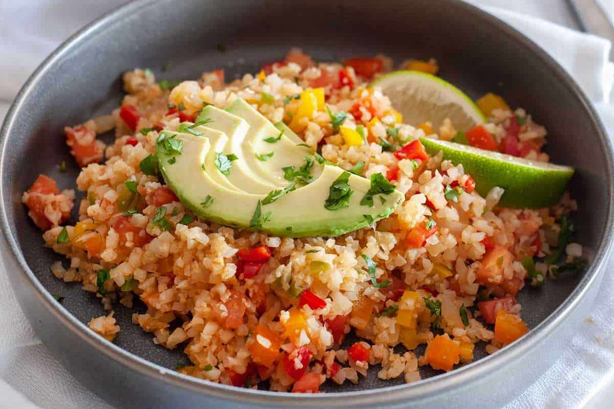 A bowl of cauliflower rice with avocado, lime, and diced bell peppers, garnished with fresh parsley.