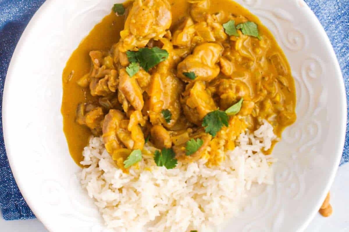 A plate of chicken curry and rice garnished with fresh cilantro, served on a white ornate plate with a blue background.