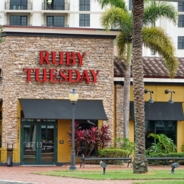 Exterior of a Ruby Tuesday restaurant offering a gluten-free menu, with a red sign, surrounded by palm trees and ornamental plants, located in front of a multi-story building.