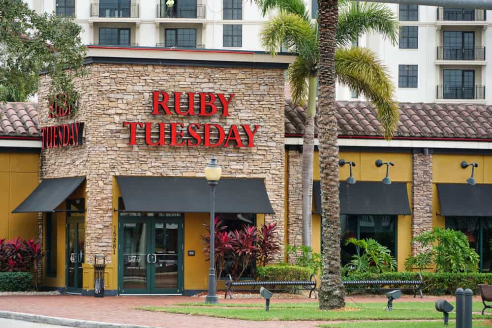 Exterior of a Ruby Tuesday restaurant offering a gluten-free menu, with a red sign, surrounded by palm trees and ornamental plants, located in front of a multi-story building.