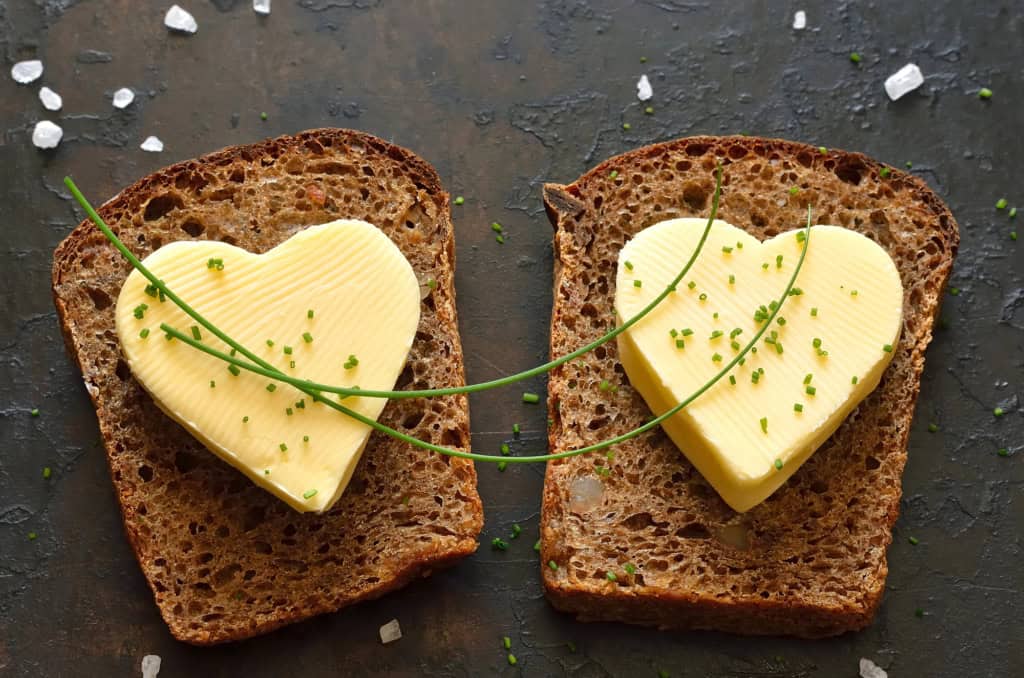 Two slices of whole grain bread topped with heart-shaped butter and garnished with chives, on a dark background with scattered salt.