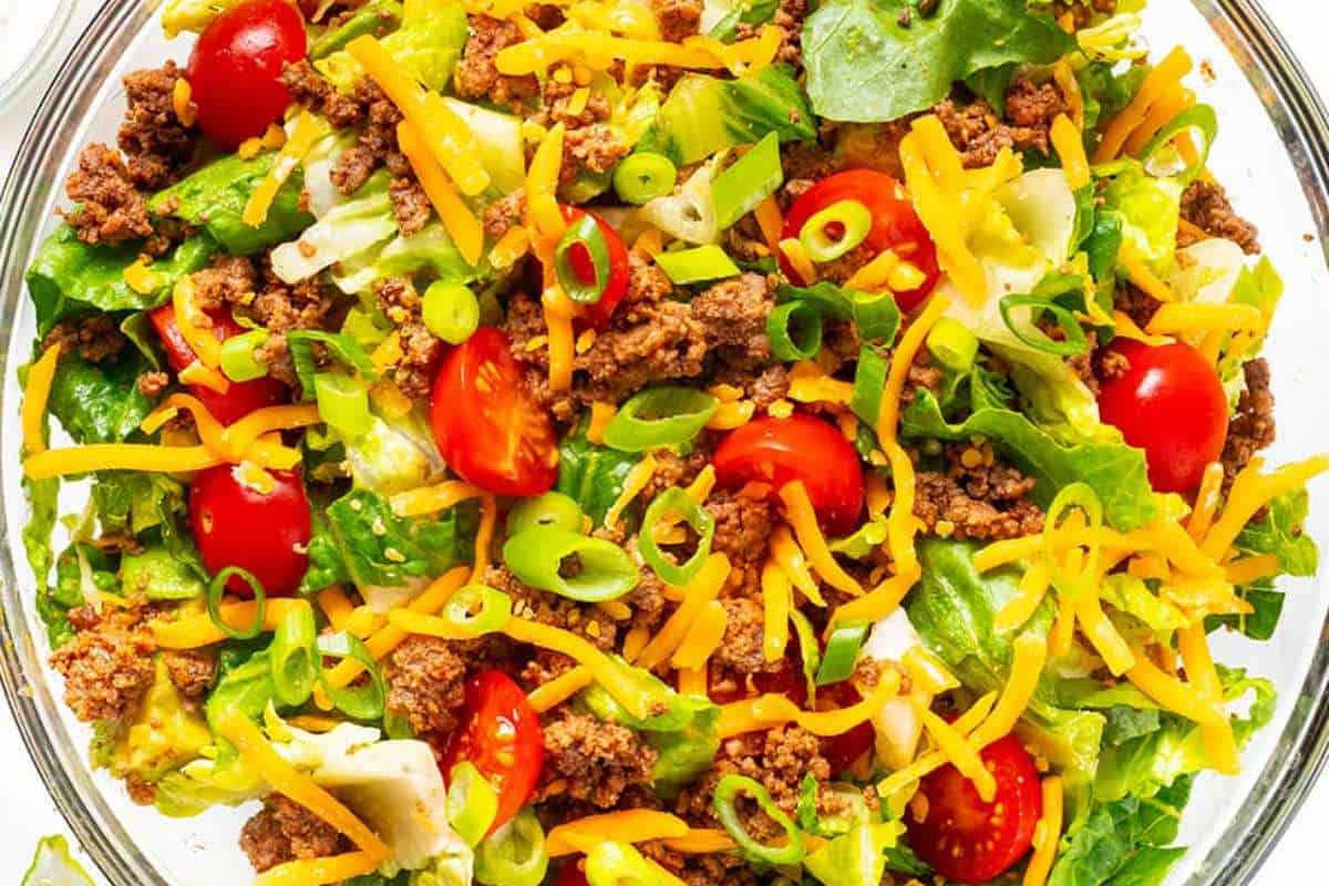 A close-up image of a taco salad featuring ground beef, shredded cheese, tomatoes, green onions, and lettuce in a glass bowl.