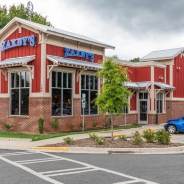 Exterior view of a Zaxby's restaurant featuring a gluten-free menu, with a red and white facade, large windows, and a parked blue car nearby under a cloudy sky.