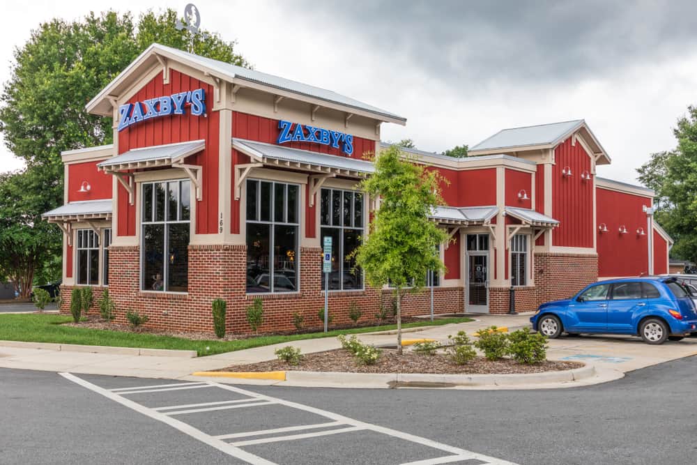 Exterior view of a Zaxby's restaurant featuring a gluten-free menu, with a red and white facade, large windows, and a parked blue car nearby under a cloudy sky.