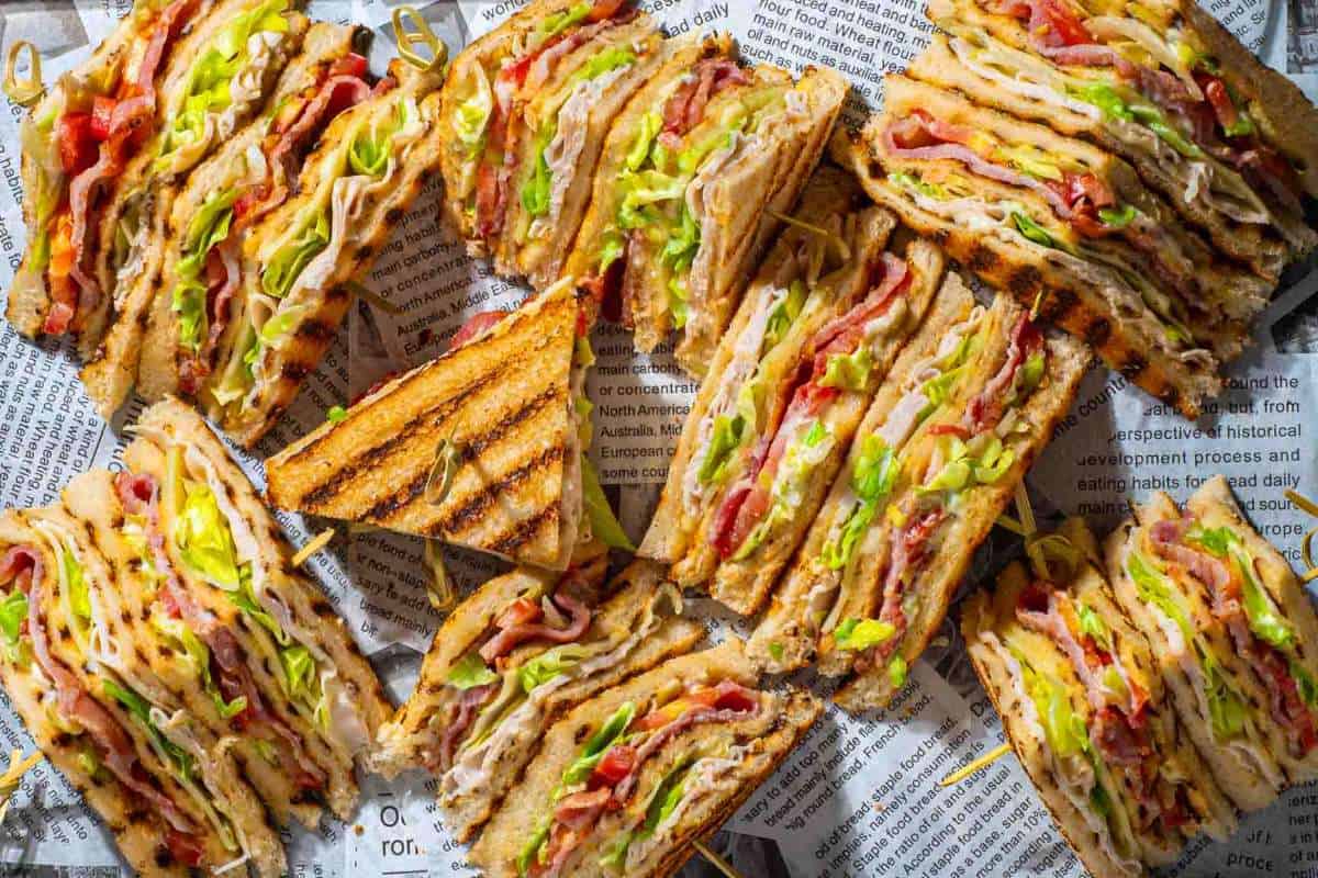 An array of toasted sandwiches filled with cheese, meat, and lettuce, cut into triangles and arranged on newspaper.