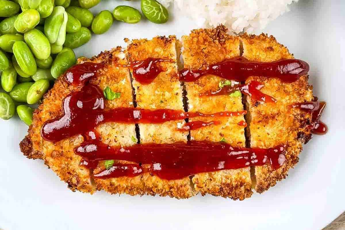 Cooked breaded chicken cutlet topped with ketchup, served with rice and green peas on a white plate.