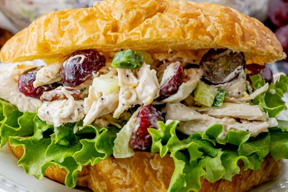 Chicken salad sandwich on a croissant with lettuce, grapes, and sliced cucumber, served on a plate.