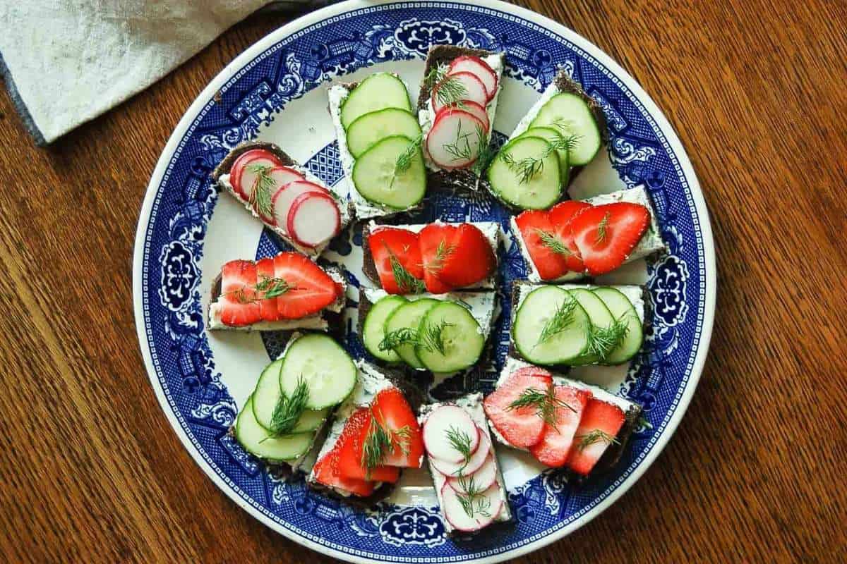 A plate with assorted open-faced sandwiches topped with cucumbers, radishes, and strawberries on a wooden table.