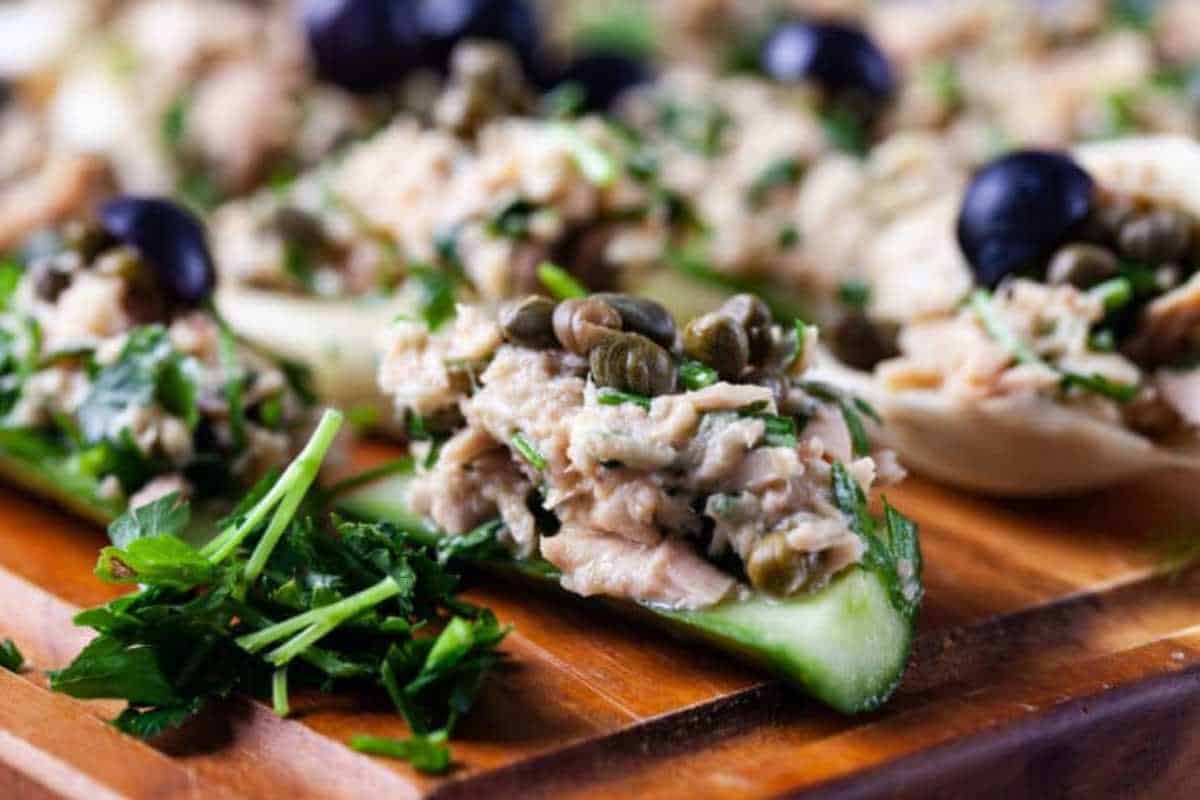 Close-up of cucumber slices topped with tuna salad, capers, and black olives, garnished with parsley on a wooden board.