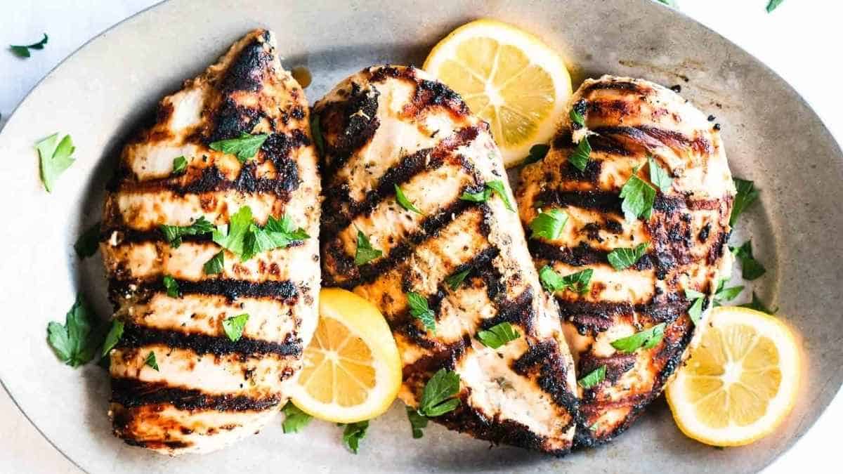 Grilled chicken breasts with char marks, garnished with parsley and lemon slices on a gray plate.