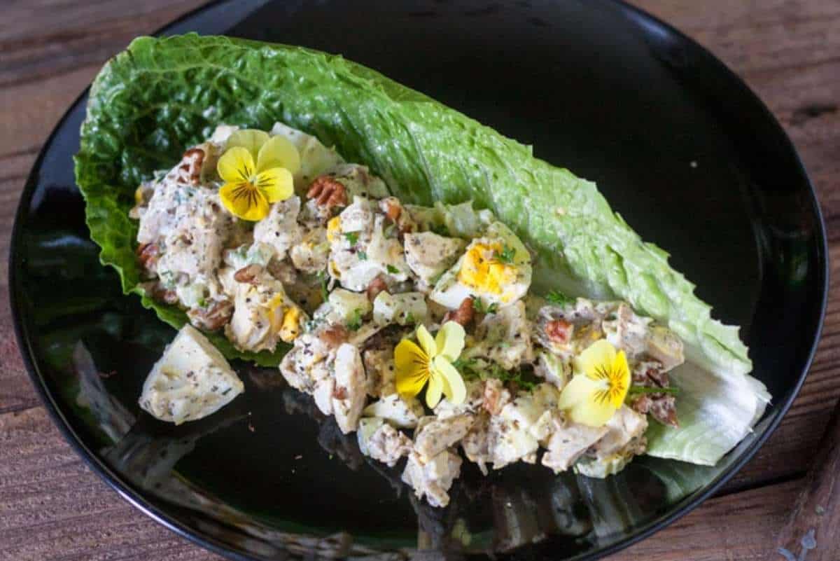 Egg salad with herbs and pecans served on a crisp romaine lettuce leaf, garnished with edible yellow flowers, presented on a black plate.