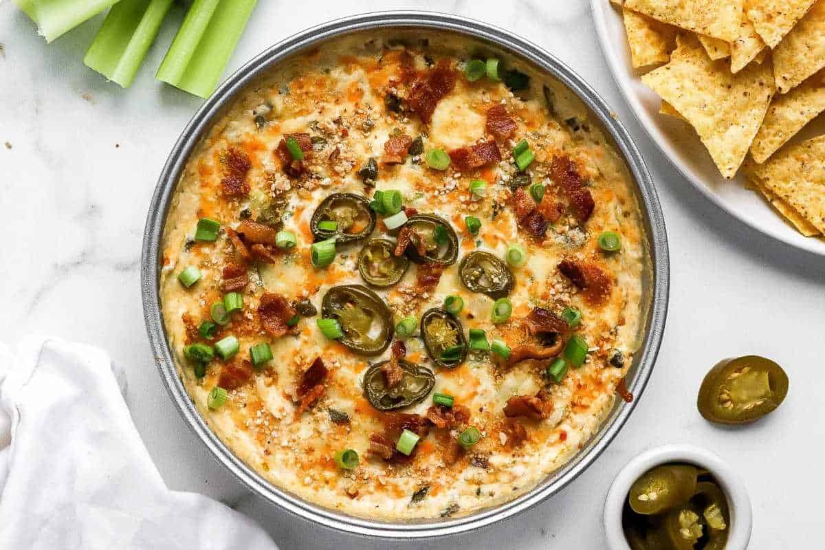 A hot serving of cheesy dip topped with bacon, jalapeños, and green onions, accompanied by a side of tortilla chips and celery sticks.