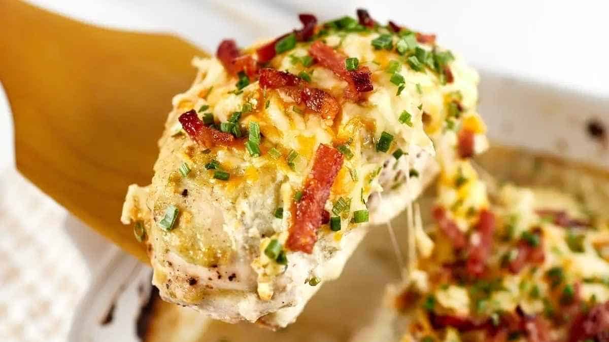 A spoon lifting a portion of creamy chicken bake topped with melted cheese, bacon bits, and chopped chives.