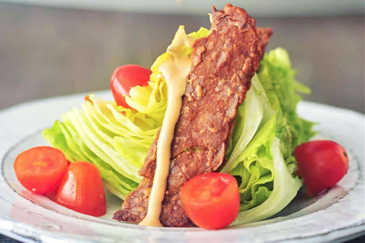 A plate of salad featuring crisp lettuce with tomatoes, topped with bacon strips and drizzled with yellow dressing.