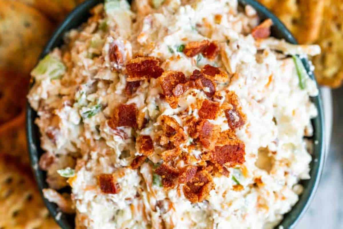 A bowl of creamy bacon dip topped with crispy bacon bits, served with crackers on the side.