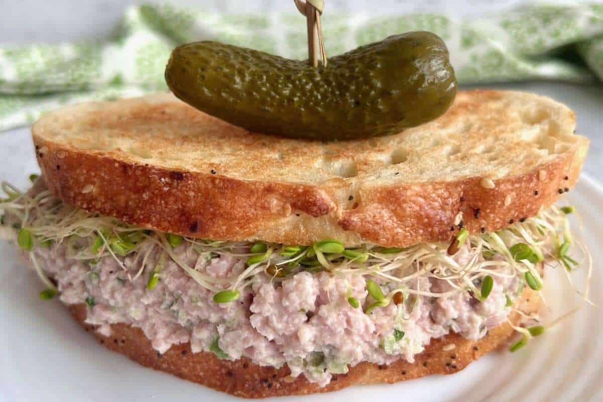 A sandwich with tuna salad and sprouts on whole grain bread, topped with a whole pickle.